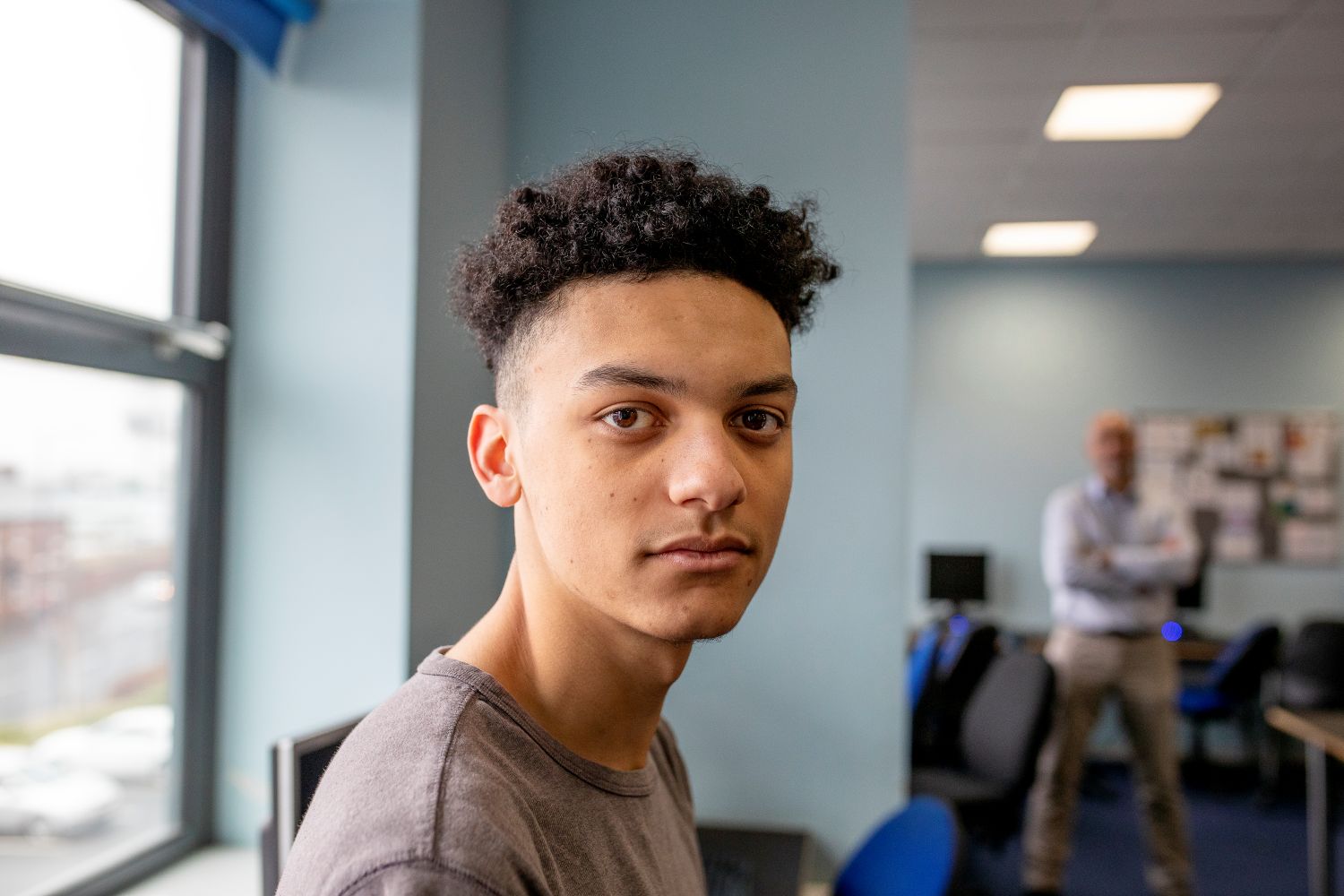 A teenage boy looks seriously at the camera - a male teacher stands behind him in the classroom.