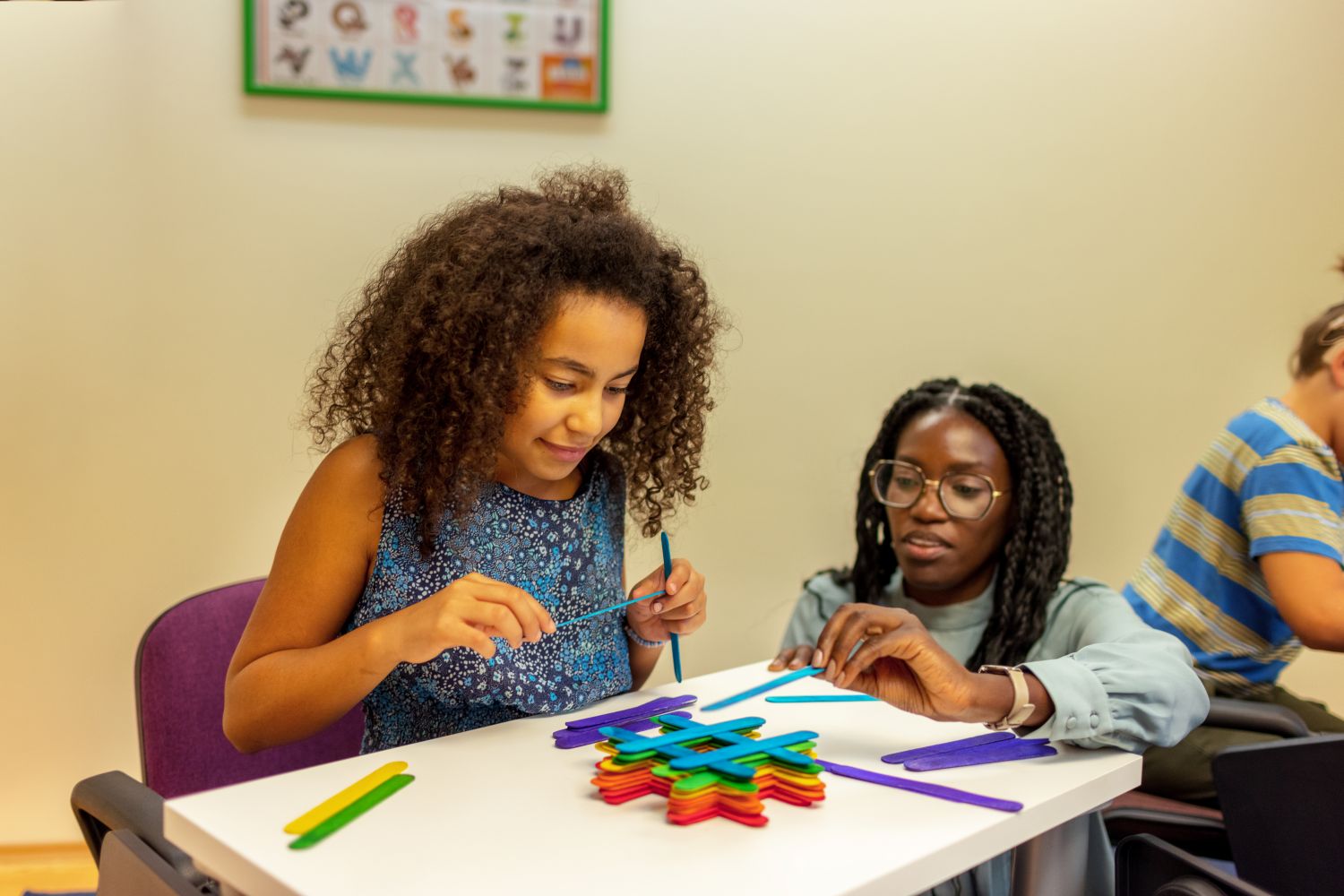 A young female teacher crouches down to help a pupil build a tower out of brightly coloured wooden lolly-pop sticks.