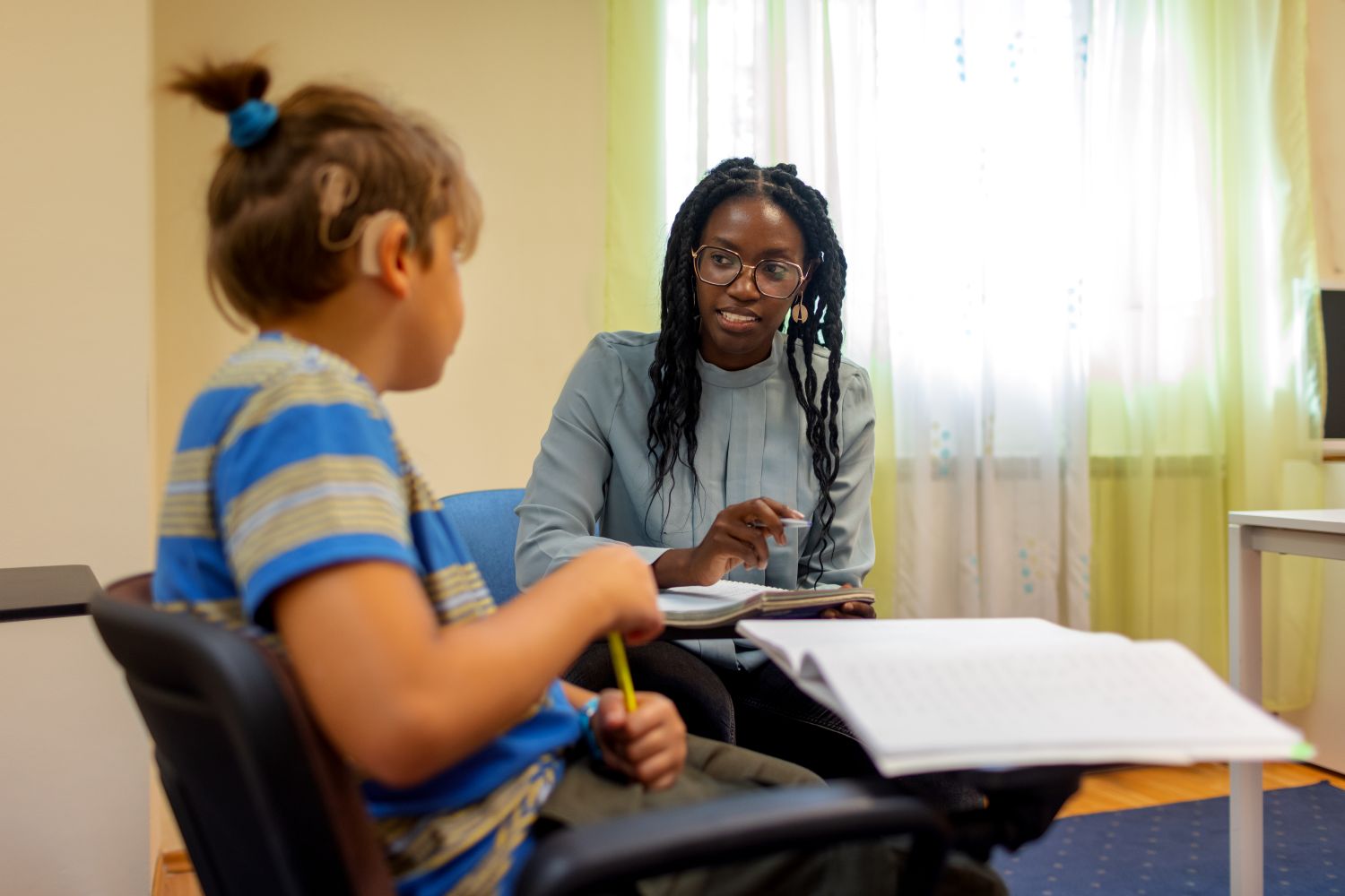 A teacher sitting in a calm room with a young deaf teenage pupil in a wheelchair, an open exercise book in front of them.
