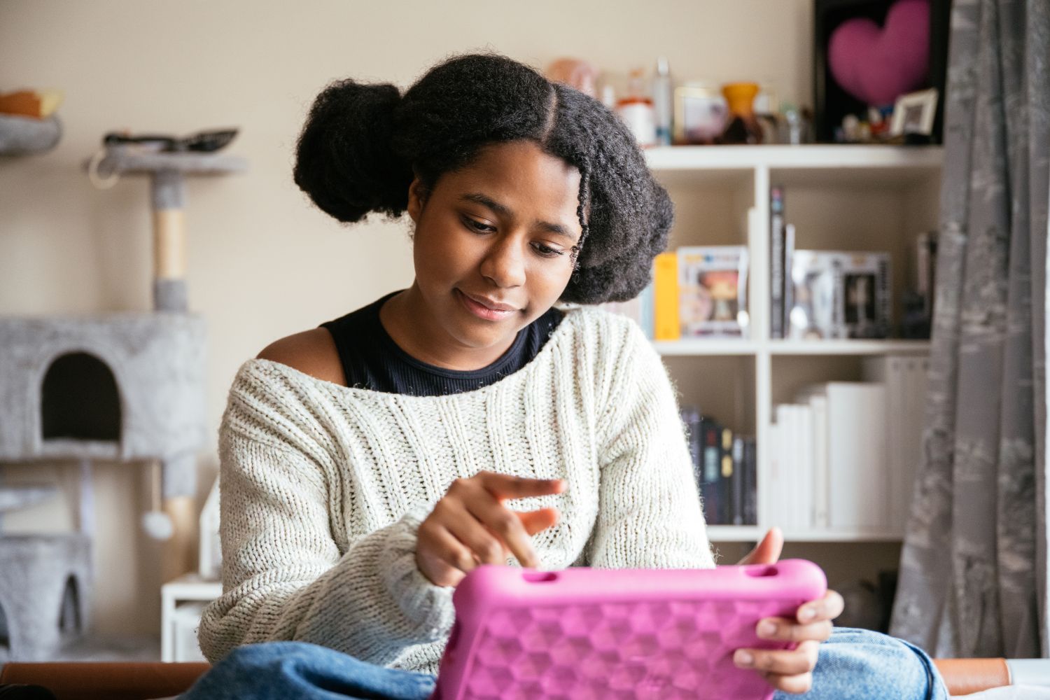 A young teenage girl with dark hair in pigtails sits cross legged whilst looking at a bright pink tablet.