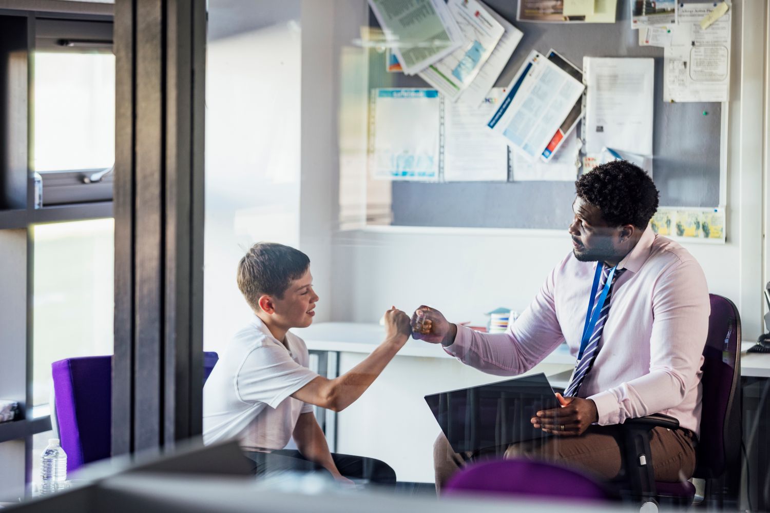 A young teenage boy and teacher bump fists in the school office.
