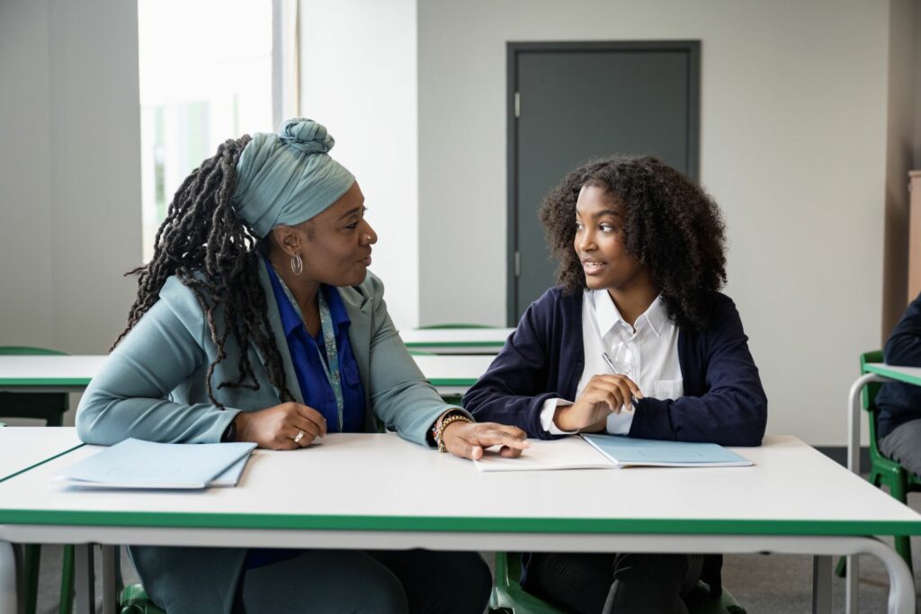 A female teacher and teenage school girl discuss work sat at a school desk. The girl is smiling as the teacher gives her gentle advice.