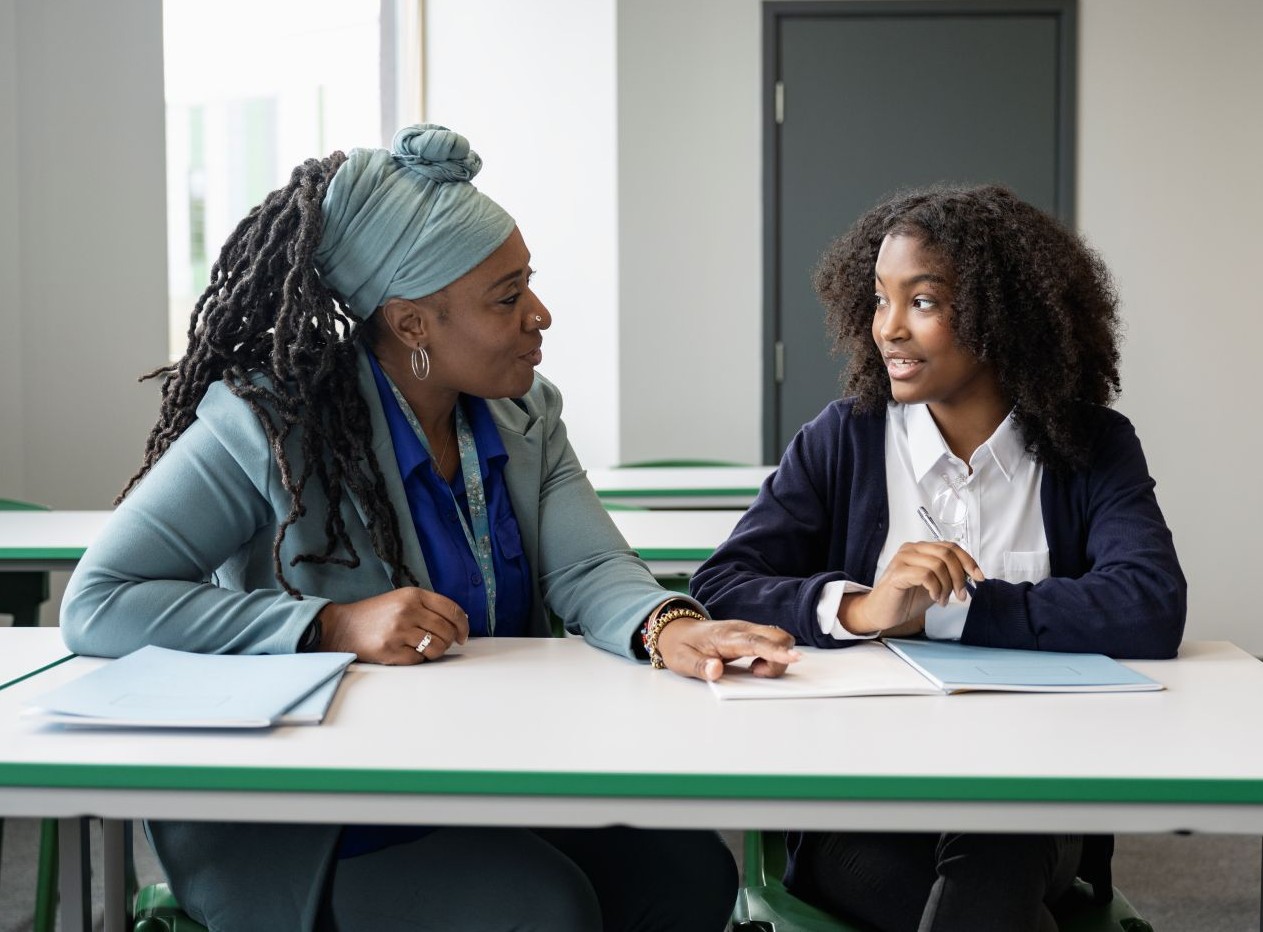 A female teacher and teenage school girl discuss work sat at a school desk. The girl is smiling as the teacher gives her gentle advice.