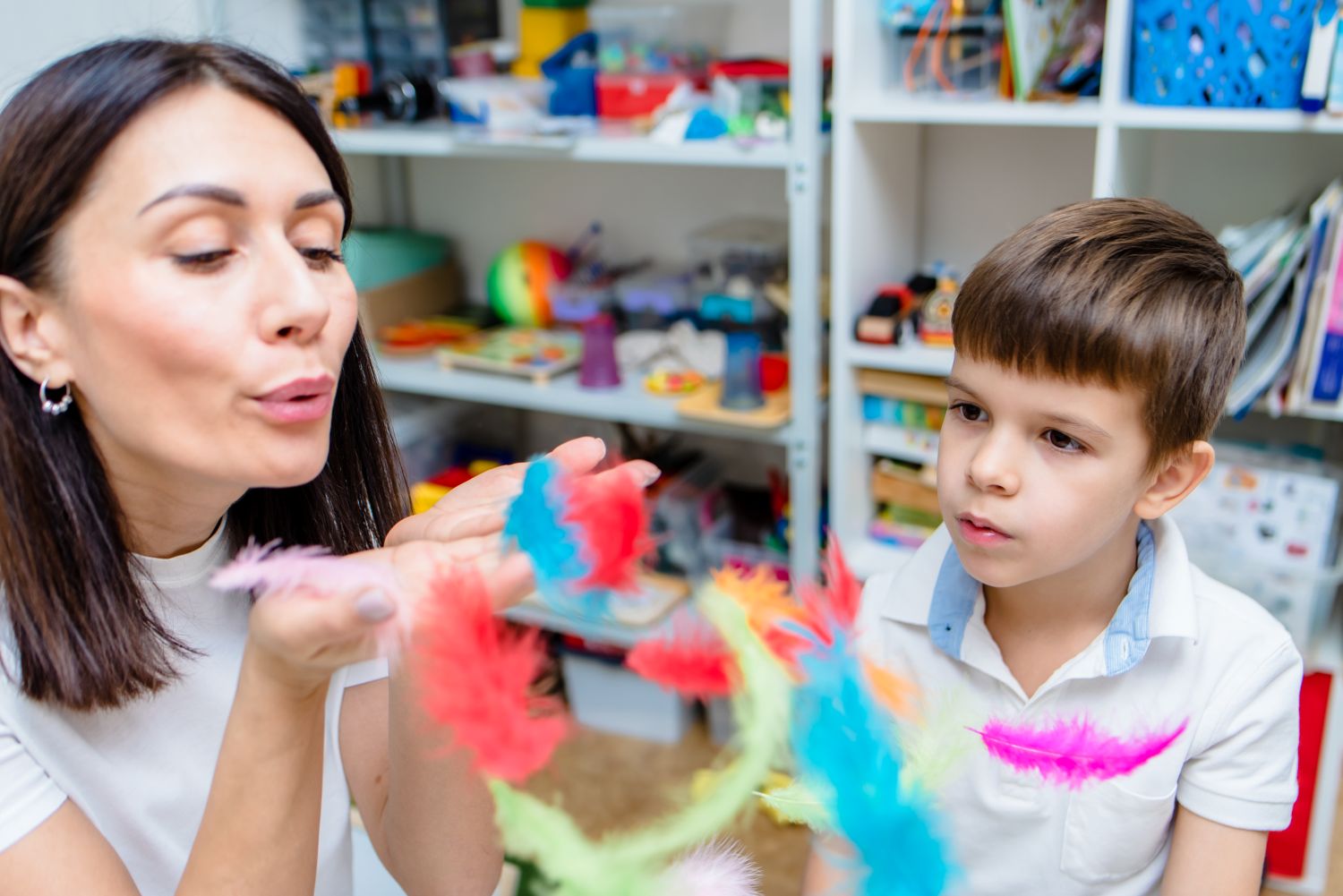 A female therapist blows a handful of brightly coloured feathers as a serious young boy watches on.