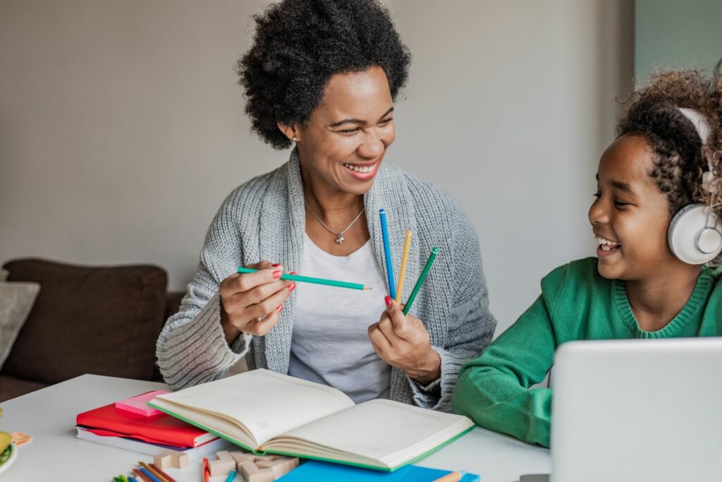 A smiling woman holding brightly coloured pencils sits next to a laughing young child wearing headphones in front of a laptop.