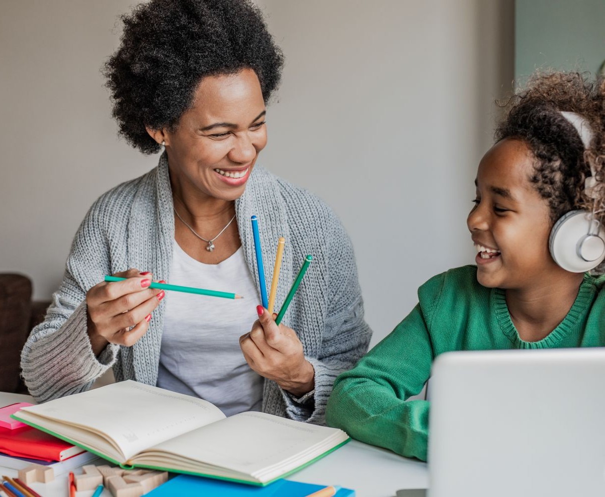 A smiling woman holding brightly coloured pencils sits next to a laughing young child wearing headphones in front of a laptop.