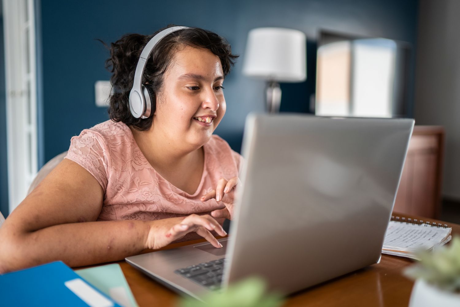 A young teenage girl with disabilities works on her laptop at home. She is wearing a pink top and headphones.