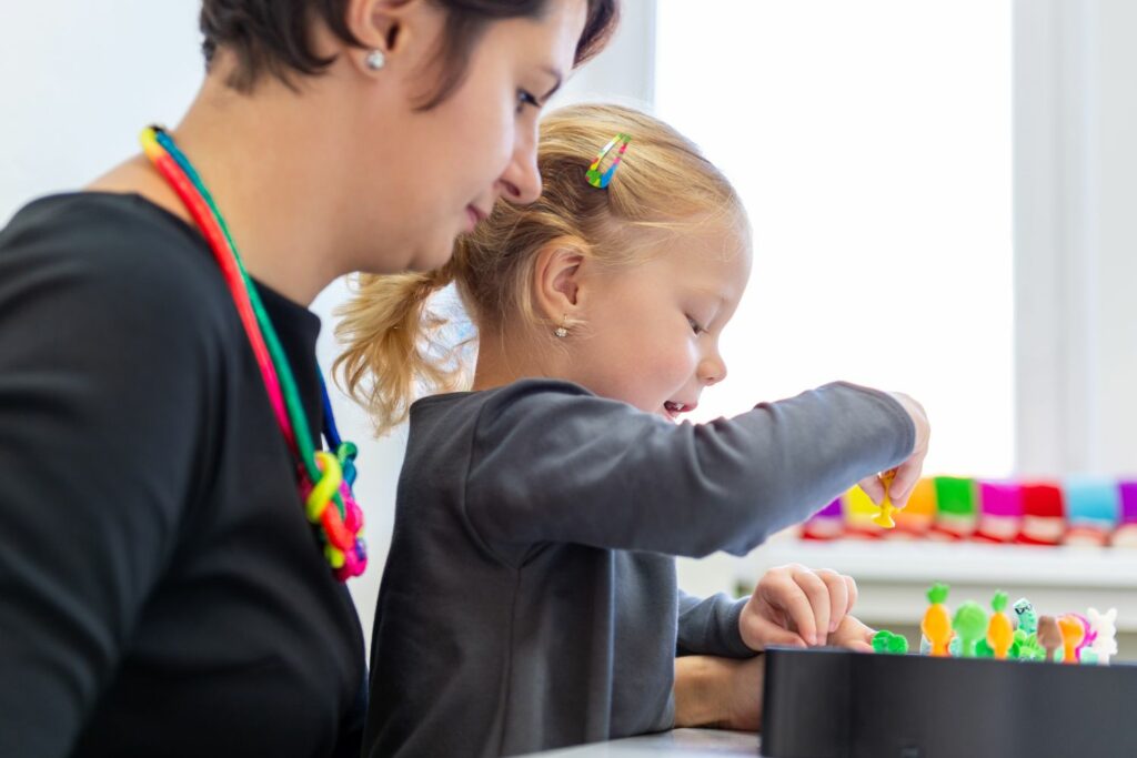 A little blonde girl plays with a vegetable activity while a female teacher watches on.