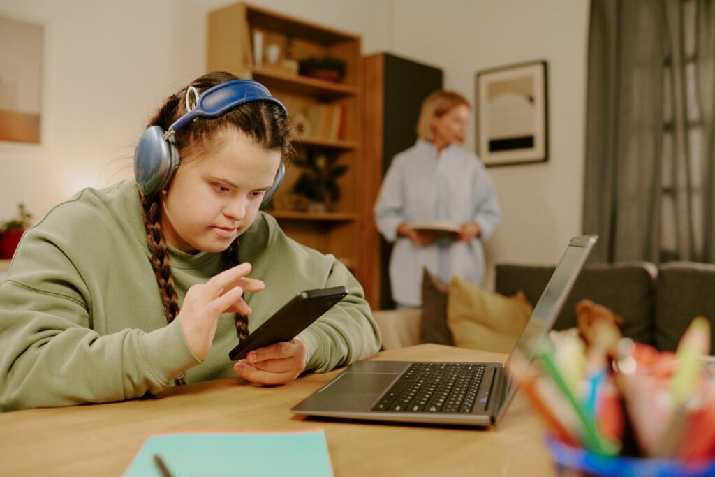 A young teenage gurl with Down's Syndrome wearing blue headphones. She is looking at her phone, with her laptop on the table in front of her, as her mum tidies in the background.