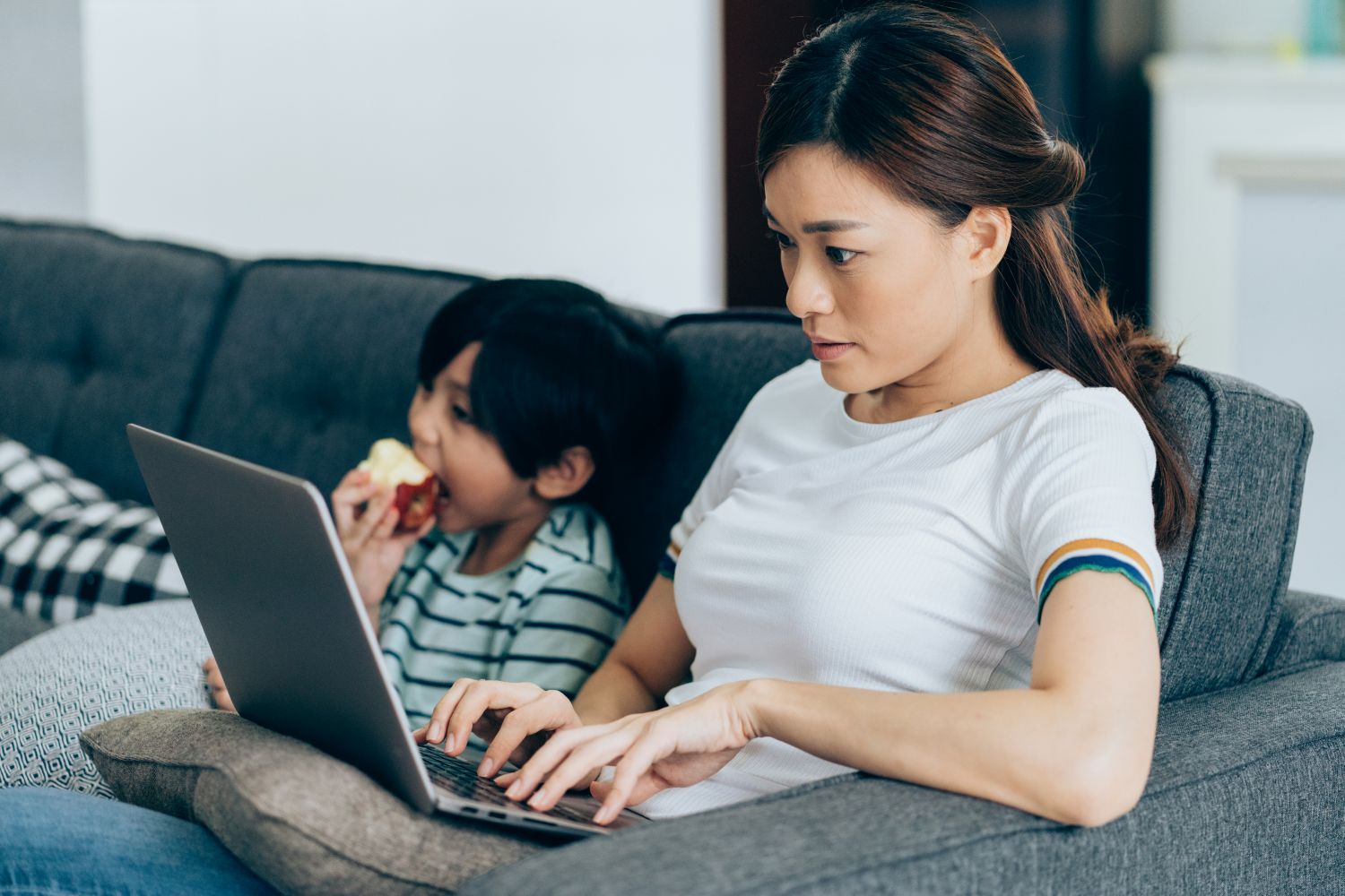 A woman with long dark hair wearing a white t-shirt with rainbow trim searches on her laptop. Next to her on the sofa is a little boy eating an apple.