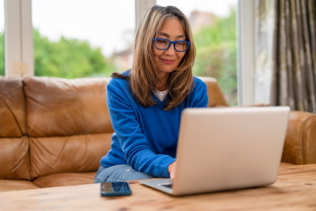 A woman in royal blue glasses and jumper sits on a brown lather sofa, working on her laptop.